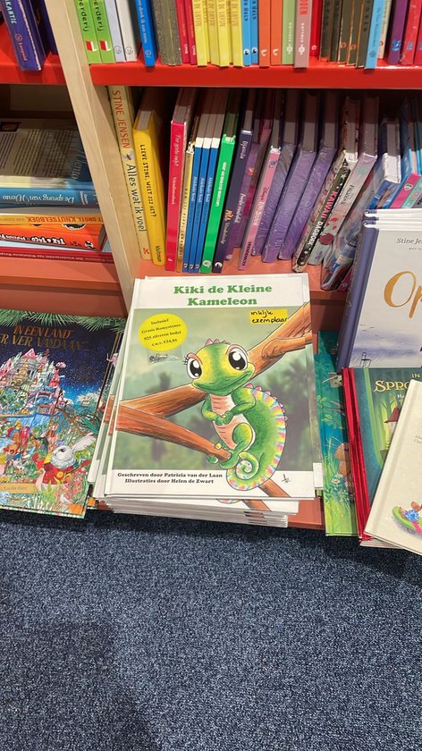 Boek "Kiki de Kleine Kameleon" op een stapel boeken in een winkel. De voorkant toont een groene kameleon met een gekleurde staart.