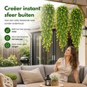 Een vrouw met een drankje, kijkt naar drie hangende kunstplanten met groene bladeren en gele bloemen op een terras.