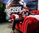 Max Verstappen op een rode stoel, gekleed in een racingsuit, met "2022 WORLD CHAMPION" op de achtergrond.