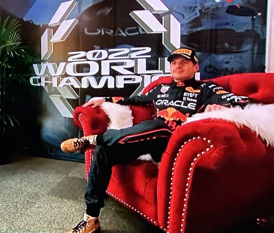 Max Verstappen op een rode stoel, gekleed in een racingsuit, met "2022 WORLD CHAMPION" op de achtergrond.