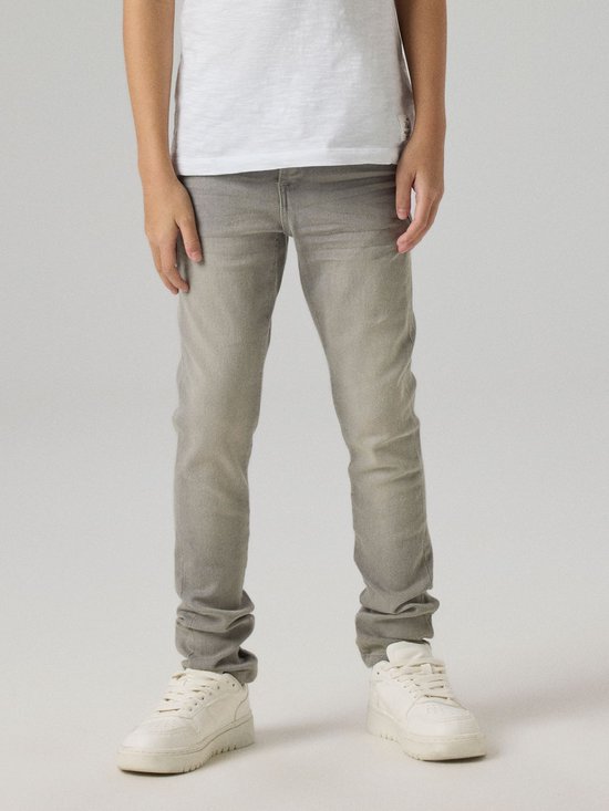 Jongen draagt een grijze, aansluitende jeans met witte sneakers en een wit t-shirt.