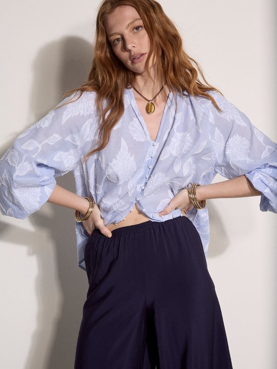 Model in losvallende blauw gestreepte blouse met bloemenpatroon en donkere ballonbroek.