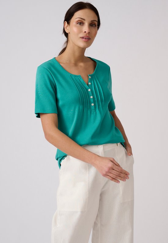 T-shirt vert pour femme, avec encolure en V et boutons, porté avec un pantalon blanc.