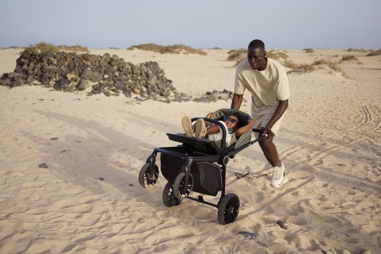Een man met een beige outfit duwt een donkergroene kinderwagen met een kind erin, in een zanderige omgeving.