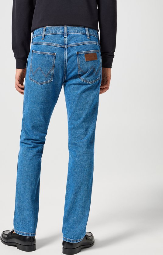 Model draagt Wrangler Greensboro jeans in 'Raindrop' wassing. Ze zijn blauw met bruin leren merklabel op de achterkant.