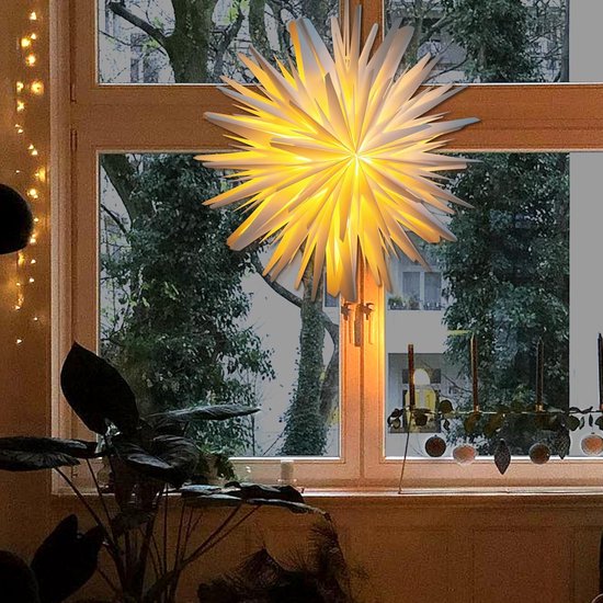 Witte papieren sterdecoratie met warm licht, hangend voor een raam met zicht op bomen.