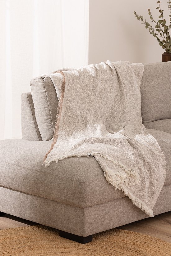 Taupe plaid met franjes gedrapeerd over de armleuning van een grijze stoffen bank. De plaid is deels uitgevouwen.