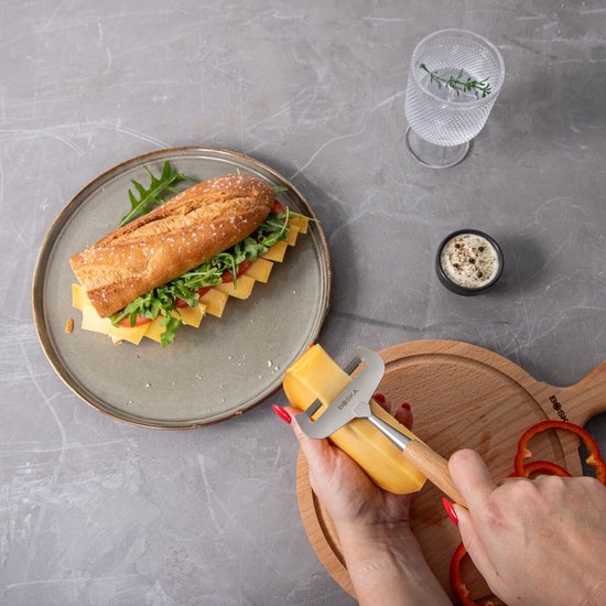 Hand snijdt kaas met een Boska kaasschaaf met houten handvat. Broodje met kaas en salade op bord.
