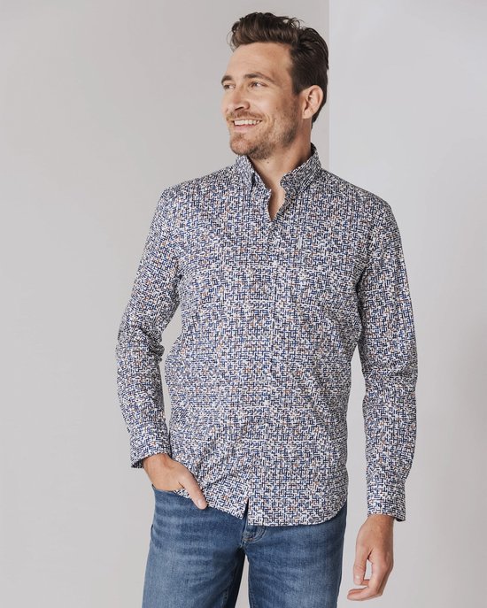 Man met hand in broekzak, draagt een blauw overhemd met een wit, bruin en donkerblauw patroon. Hij kijkt weg.
