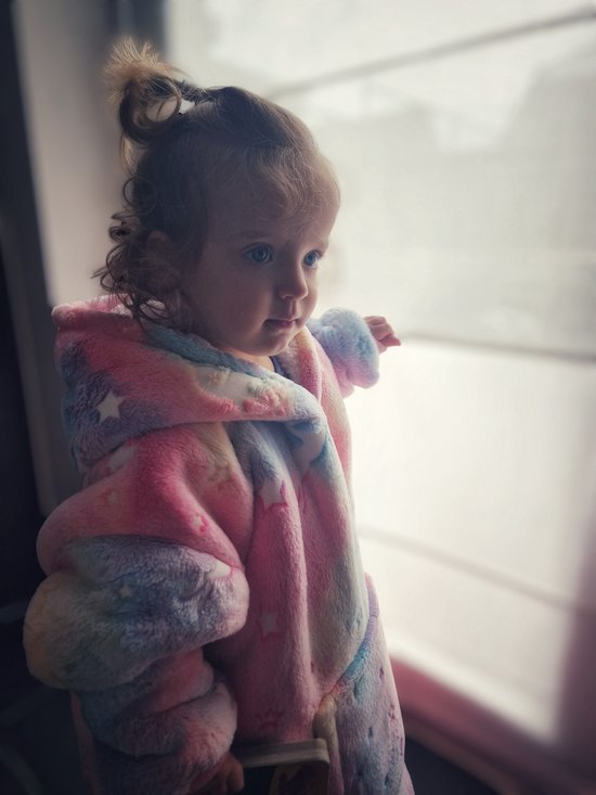 Jong meisje met krullend haar, staand bij een raam, draagt een pluche hoodie deken met pastel regenboogkleuren, sterren en eenhoorns.