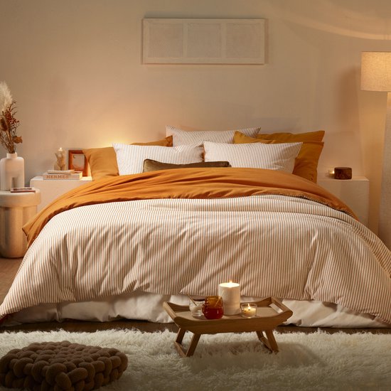Un lit fait avec une housse de couette couleur crème à fines rayures brunes, une couverture orange et des coussins.