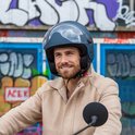 Man met donkerblauwe helm met vizier en beige jas, lachend, zittend op een scooter.