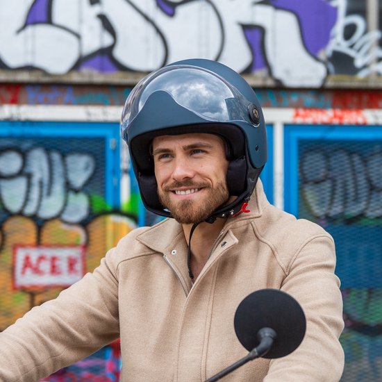 Man met donkerblauwe helm met vizier en beige jas, lachend, zittend op een scooter.
