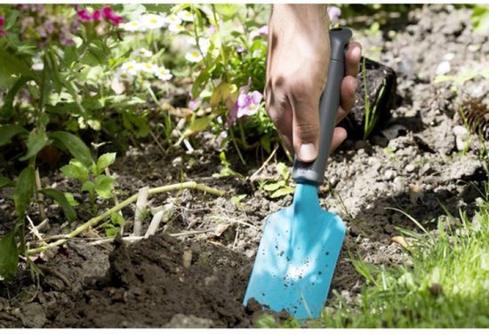 Een hand houdt een blauw tuinschepje met grijs handvat vast, terwijl het in de aarde wordt gedrukt tussen planten.