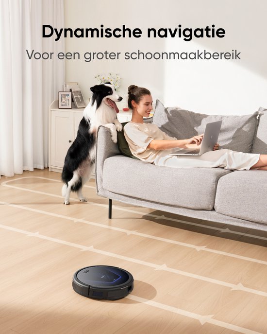 Zwarte eufy robotstofzuiger volgt witte pijlen op houten vloer. Vrouw zit op bank met hond en laptop.