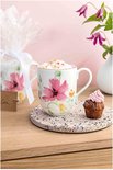 Witte mok met roze bloemenprint, gevuld met dranken en bestrooid met hagelslag, naast een cupcake.