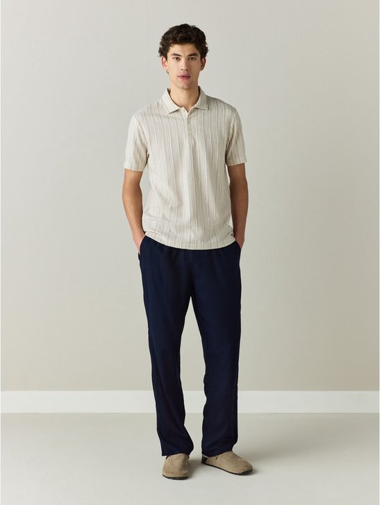 Volledige outfit van een man in een beige polo met fijne verticale strepen en een marineblauwe broek, beige schoenen.
