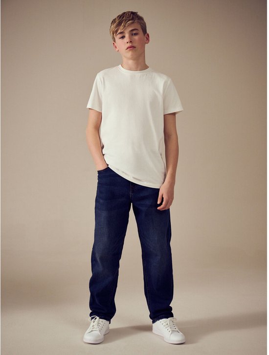 Jongen met blonde haren draagt een donkerblauwe spijkerbroek en wit T-shirt.