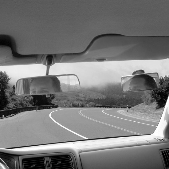 Reflectie van een gezicht in twee autospiegels, met een bochtige weg en mist op de achtergrond.