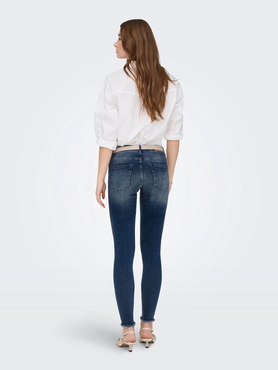 Vrouw met lang bruin haar in een witte blouse en donkerblauwe skinny jeans met rafels aan de onderkant.