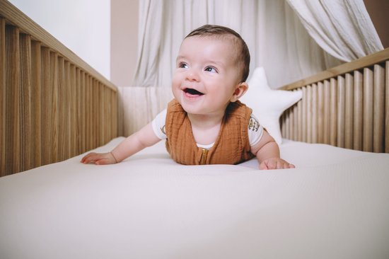 Vrolijke baby liggend op een witte, zachte matras in een houten ledikant.