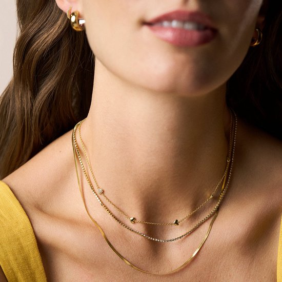 Drie gouden kettinkjes gedragen door een vrouw: een met hartjes en steentjes, een met veel kleine steentjes en een platte slangenhuid-look.