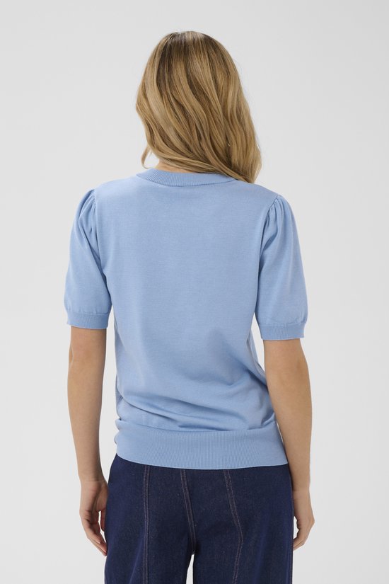 Achterkant van een blonde vrouw in een lichtblauwe trui met korte, gepofte mouwen en een donkere spijkerbroek.