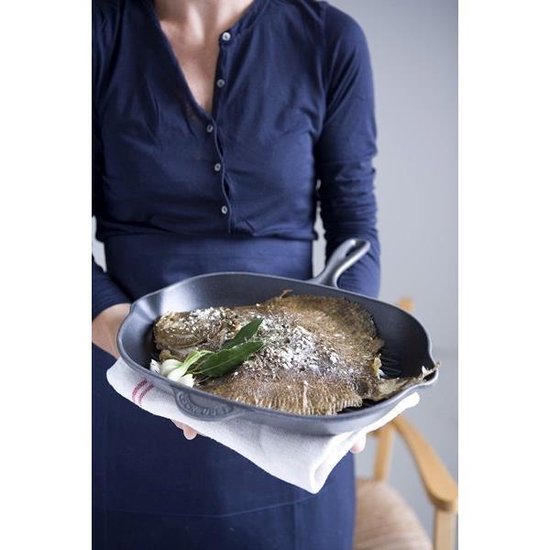 Vrouw in donkerblauw shirt houdt een zwarte gietijzeren pan met een gegrilde vis vast.