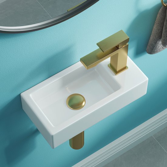 Lavabo en céramique blanc avec robinet doré, bouchon de vidange doré et siphon doré fixé au mur.