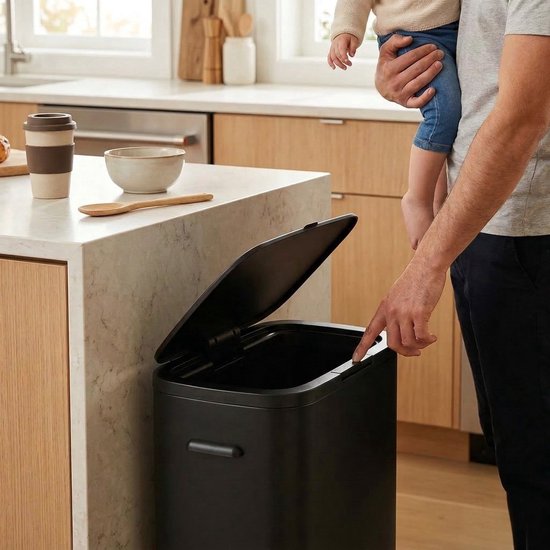 Een man met baby op armen opent zwarte prullenbak met zijn vinger in een keuken.