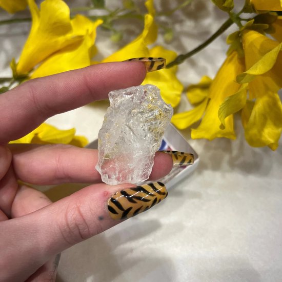 Een helder, ruw bergkristal vastgehouden tussen een hand met tijgerprint nagels en gele bloemen.