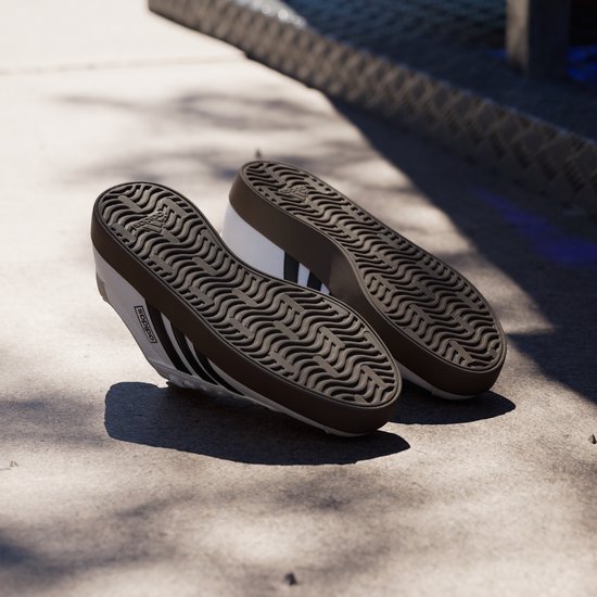 Paar witte Adidas sneakers met zwarte strepen, bruine zolen, op een betonnen ondergrond.
