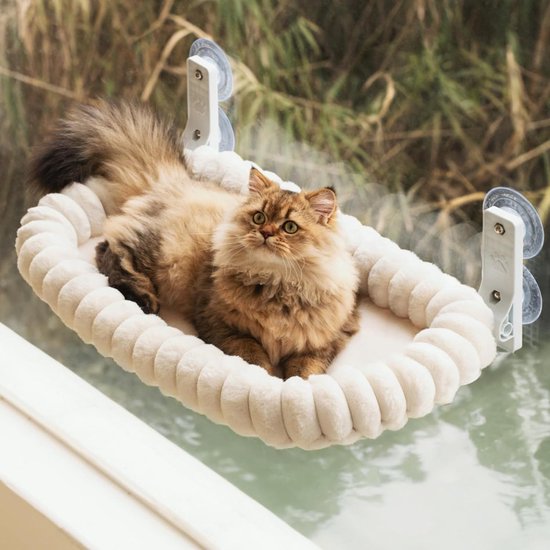 Een langharige kat met goudkleurige ogen ligt op een zacht, crèmegekleurd kattenbed met geribbelde randen, bevestigd aan een raam.