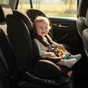 Baby met blonde haren in een zwart autostoeltje met gestructureerde rugleuning en hoofdsteun. De baby houdt een gekleurd speeltje vast.