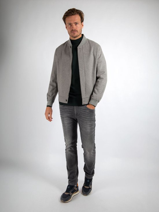 Man in grijze bomberjack met donkergroene coltrui en jeans, staand.