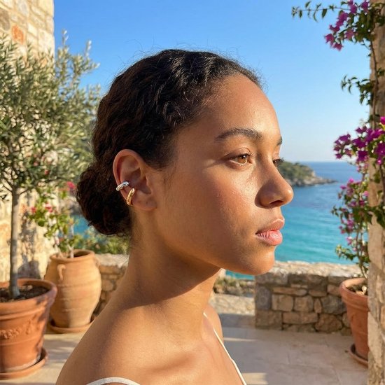 Een jonge vrouw met zilverkleurige en goudkleurige ear cuffs aan één oor, met de zee op de achtergrond.