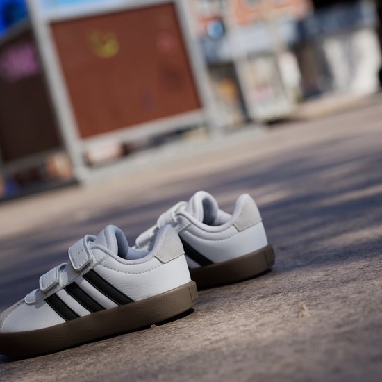 Twee witte sneakers met zwarte strepen, klittenbandsluiting en bruine zool op een stenen ondergrond.