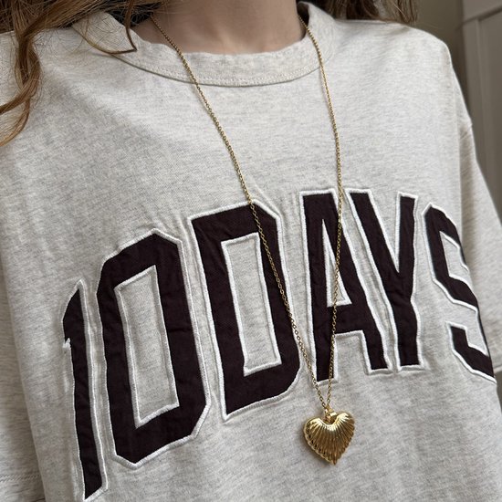 Een gouden hartvormige ketting met een reliëfpatroon, gedragen over een grijze trui met de tekst '100 DAYS'.