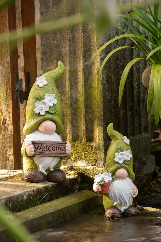 Deux nains verts avec des fleurs sur leur chapeau, debout dans un jardin. L'un tient un panneau de bienvenue.