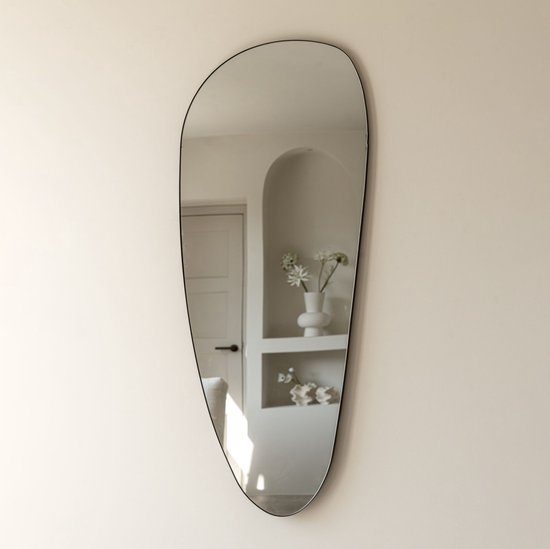 Asymmetrische zwarte spiegel met een slanke lijst, hangend aan een lichte muur. In de spiegel is een halfronde nis te zien met een witte vaas en bloemen.