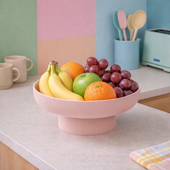 Roze fruitschaal op voet gevuld met bananen, appels, sinaasappels en druiven, op een aanrecht.