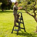 Man in groene shirt snoeit een boom met een schaar, staande op een zwarte ladder in een grasveld.