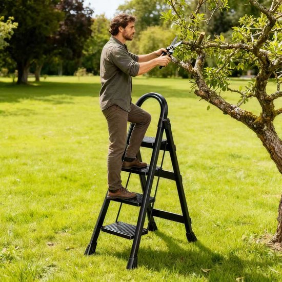 Man in groene shirt snoeit een boom met een schaar, staande op een zwarte ladder in een grasveld.