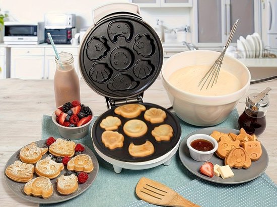 Een wafelijzer met open deksel, met beslag en diverse vormen wafels op een tafel.