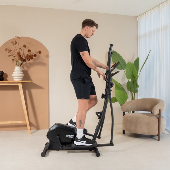 Man op een zwarte crosstrainer, in een woonkamer, voor een vaas met droogbloemen.