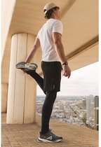 Man strekt zijn been in donkerblauwe Allrounder sneakers, zwarte sportbroek en witte T-shirt. Stadslandschap op de achtergrond.