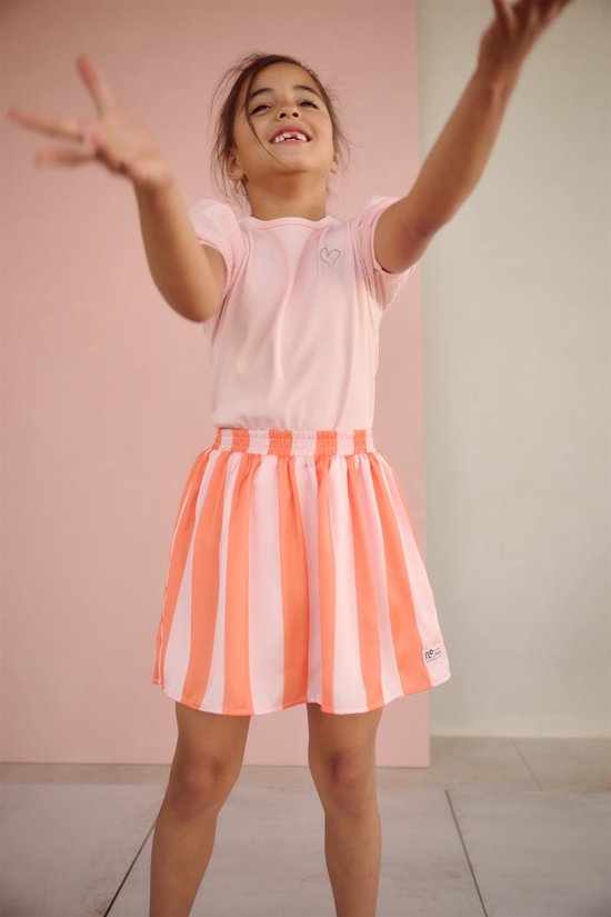 Jong meisje met haar armen omhoog, lachend, met een lichtroze t-shirt met glitterhartje en een oranje-roze gestreepte rok.