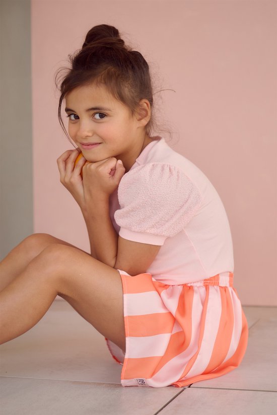 Jong meisje met een hoge knot, zittend, met handen bij de kin, met een lichtroze t-shirt met glitterhartje en een oranje-roze gestreepte rok.
