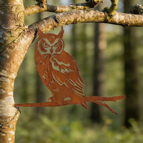 Roestbruine metalen uil van cortenstaal, zittend op een tak in een bosrijke omgeving.