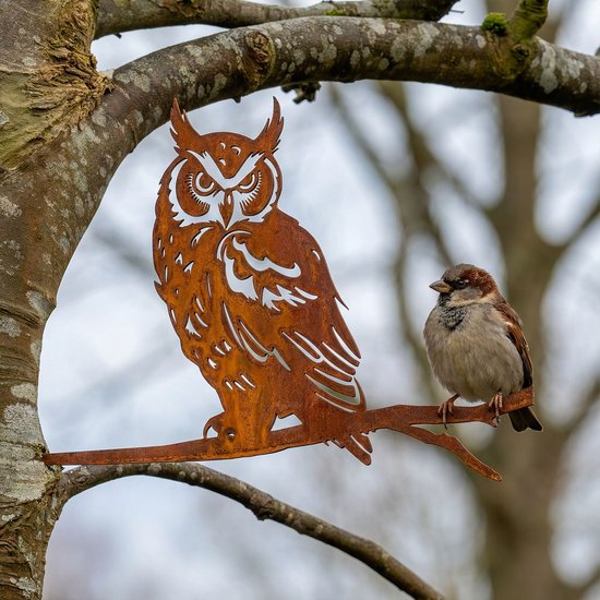 Roestbruine metalen uil van cortenstaal, naast een kleine vogel op een tak.